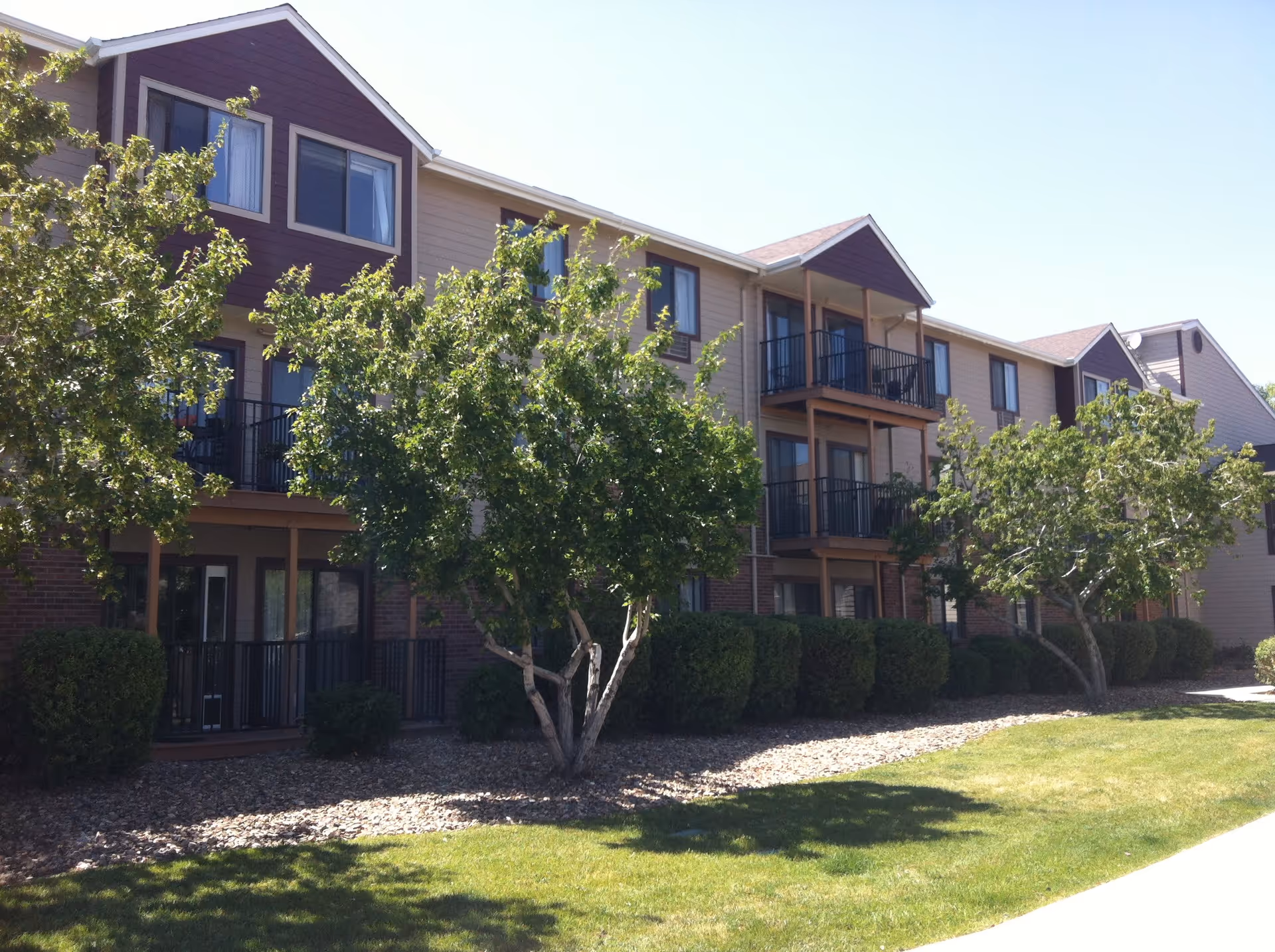 Exterior view of a three-story senior living facility building with balconies, surrounded by green trees, bushes, and a well-maintained lawn under a clear sky.