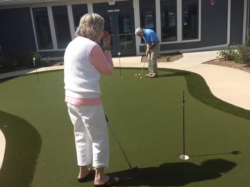 Two elderly individuals playing putting golf on a small artificial green outside a building with large windows and a door.