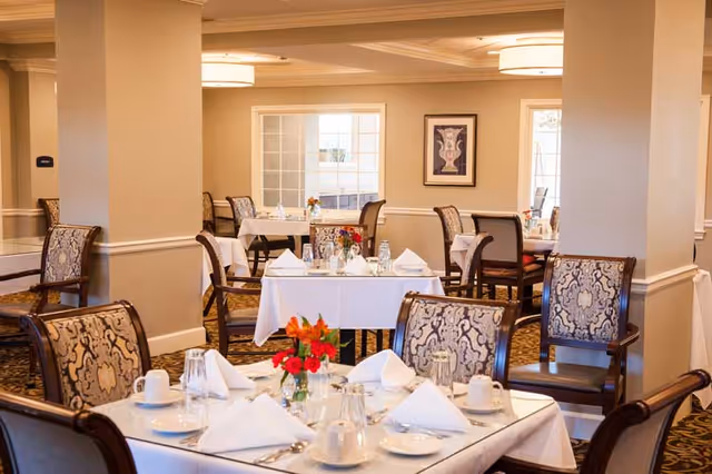A well-lit dining room with multiple tables set for a meal. Each table is covered with a white tablecloth and neatly arranged with white napkins, cups, glasses, and silverware. The chairs have patterned upholstery, and there are small floral centerpieces on the tables. The room has beige walls, large windows, and framed artwork on the walls.