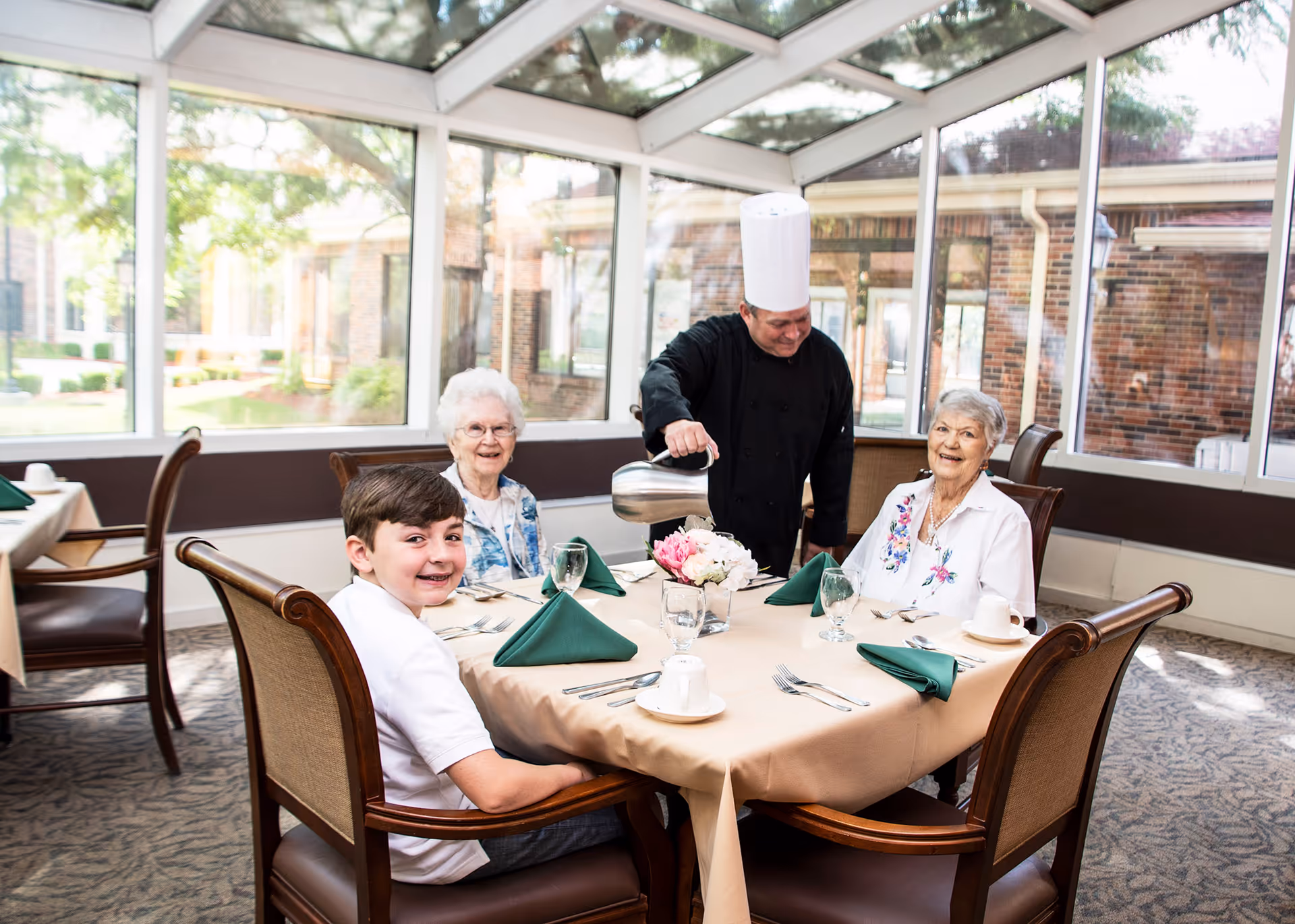 A chef in a black uniform and tall white hat is pouring water into a glass at a dining table where two elderly women and a young boy are seated. The table is set with green napkins, white cups, and silverware, and there is a floral centerpiece. The room has large windows and a glass ceiling, allowing natural light to fill the space.