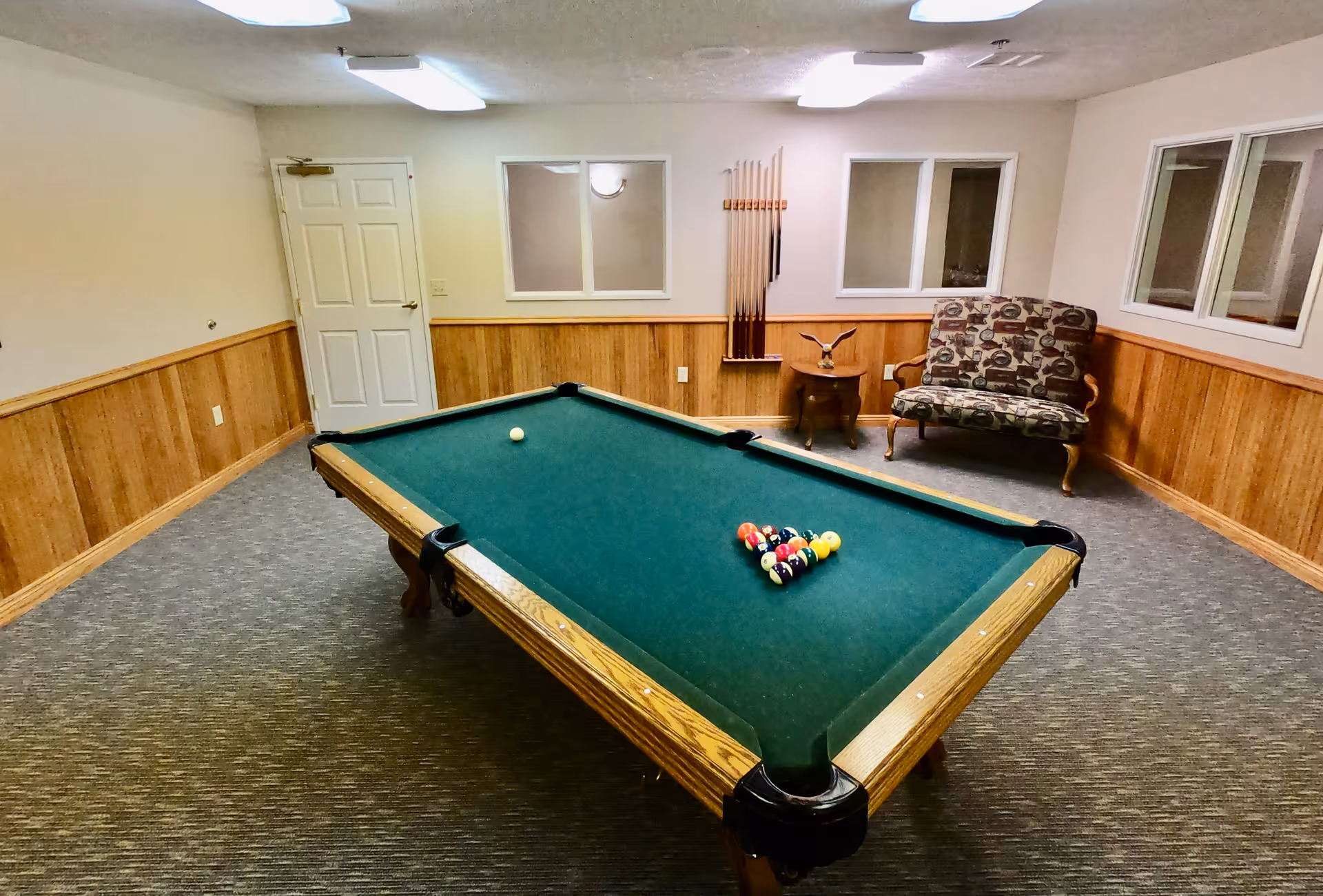 A room with a pool table set up for a game, featuring a green felt surface and a rack of billiard balls arranged in a triangle. The room has wood paneling on the lower half of the walls, beige upper walls, and carpeted floor. There is a patterned upholstered chair and a small wooden side table with a decorative eagle figurine in the corner. Pool cues are mounted on the wall behind the chair. The room is lit by ceiling fluorescent lights and has several windows.