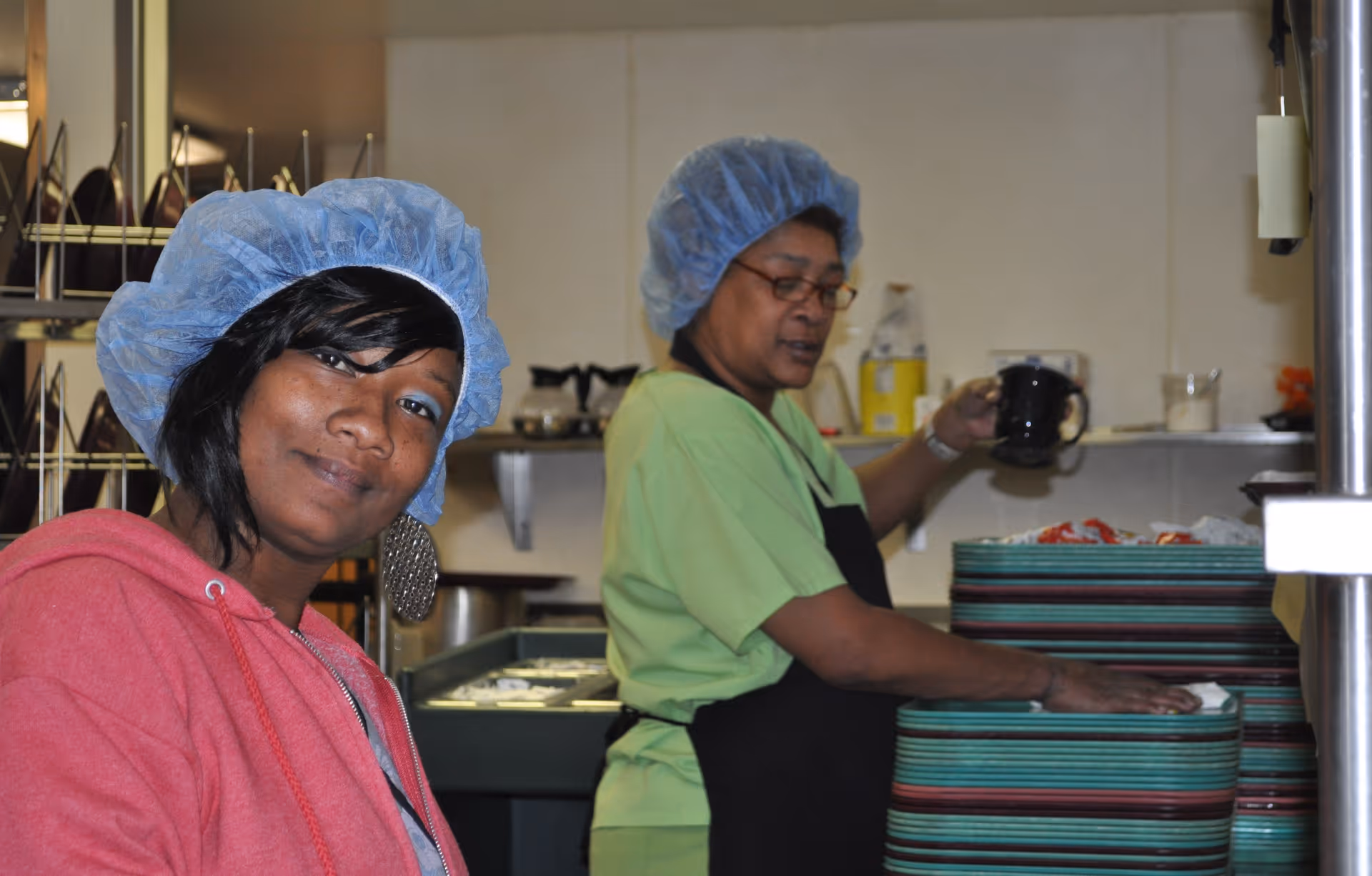 Two kitchen staff wearing hairnets stand by a stack of meal trays, one looking at the camera.
