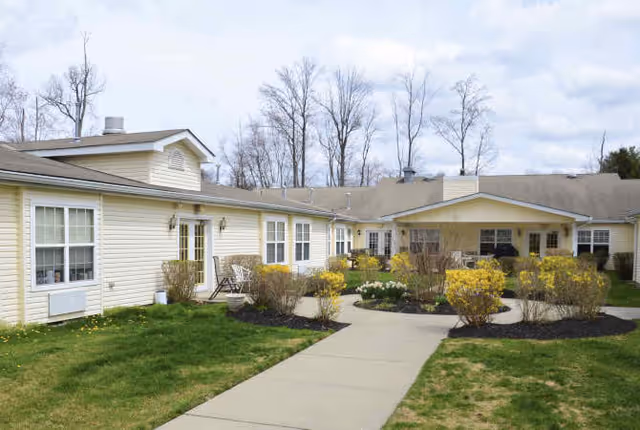 Exterior view of Granville Place, a single-story building with beige siding and multiple windows. A concrete walkway leads through landscaped bushes and flower beds towards the entrance under a covered porch. Leafless trees and a cloudy sky are visible in the background.
