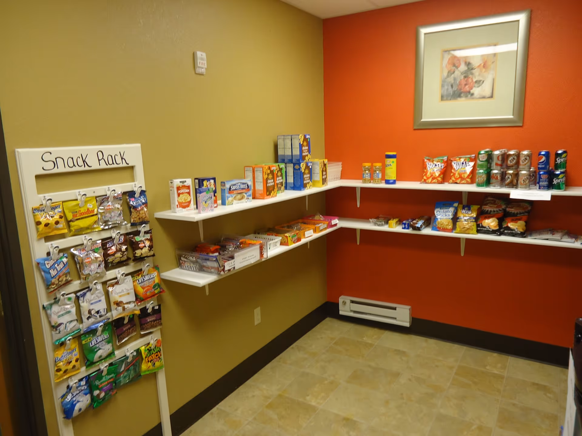 Small snack room with a 'Snack Rack' display and wall shelves stocked with chips, canned drinks and packaged snacks against red and tan walls.