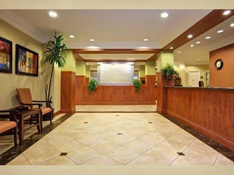 A spacious and well-lit reception area with a tiled floor, wooden paneling on the walls, and a front desk on the right side. There are chairs and framed artwork on the left wall, along with potted plants adding greenery to the space. The ceiling has recessed lighting.