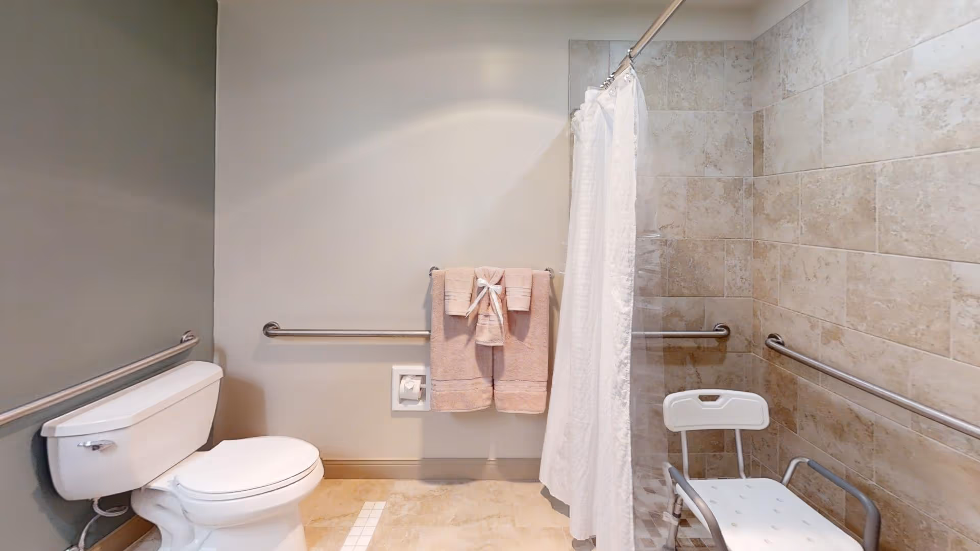 A bathroom with a white toilet on the left, beige tiled walls around a shower area on the right, a white shower curtain, and a white shower chair inside the shower. There are metal grab bars along the walls and two beige towels hanging on a towel rack in the center.