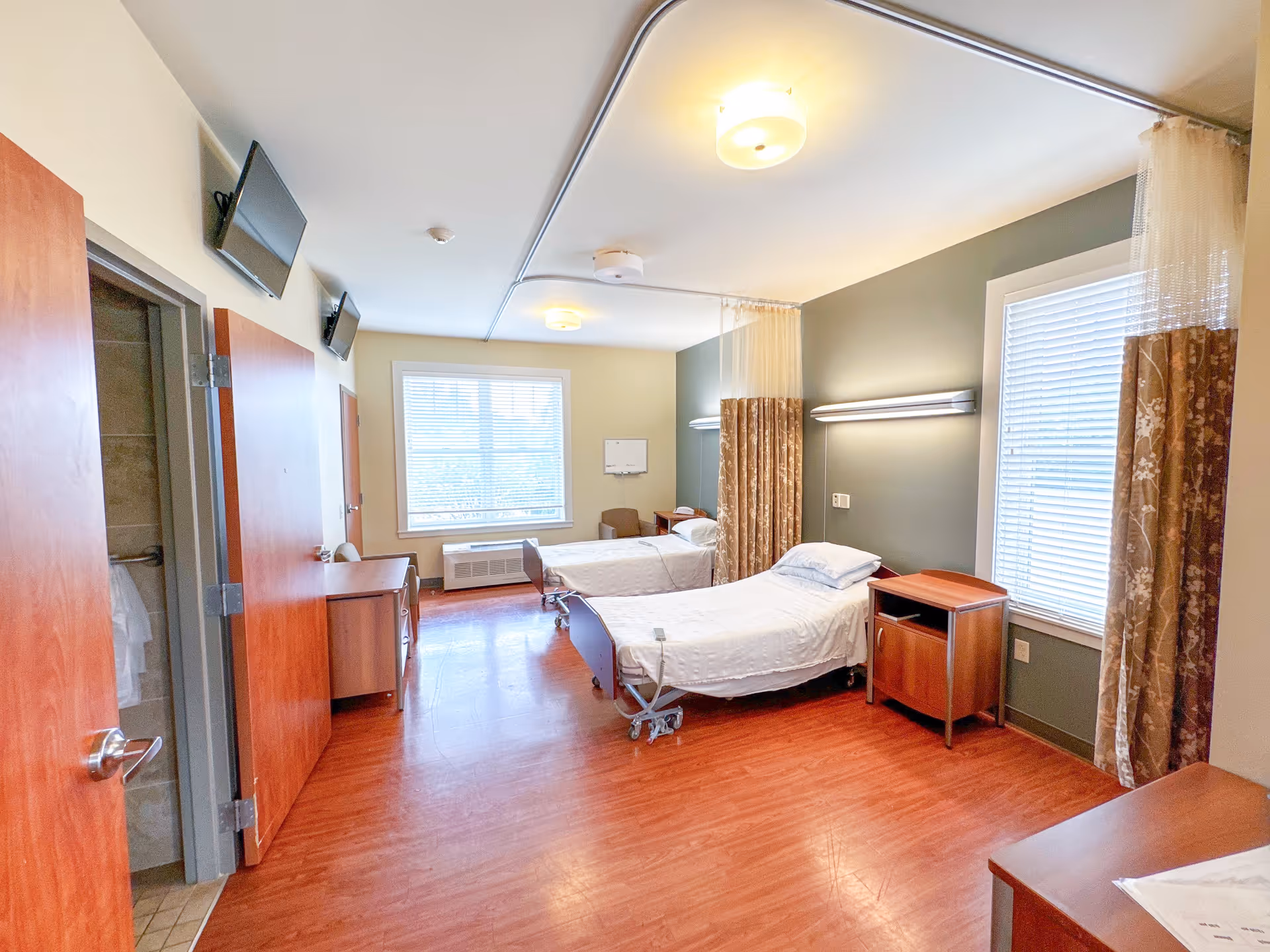 A bright and clean room with two hospital-style beds separated by a privacy curtain. Each bed has white linens and a wooden nightstand next to it. The room has wooden floors, two windows with blinds, two wall-mounted televisions, and a door leading to a bathroom.