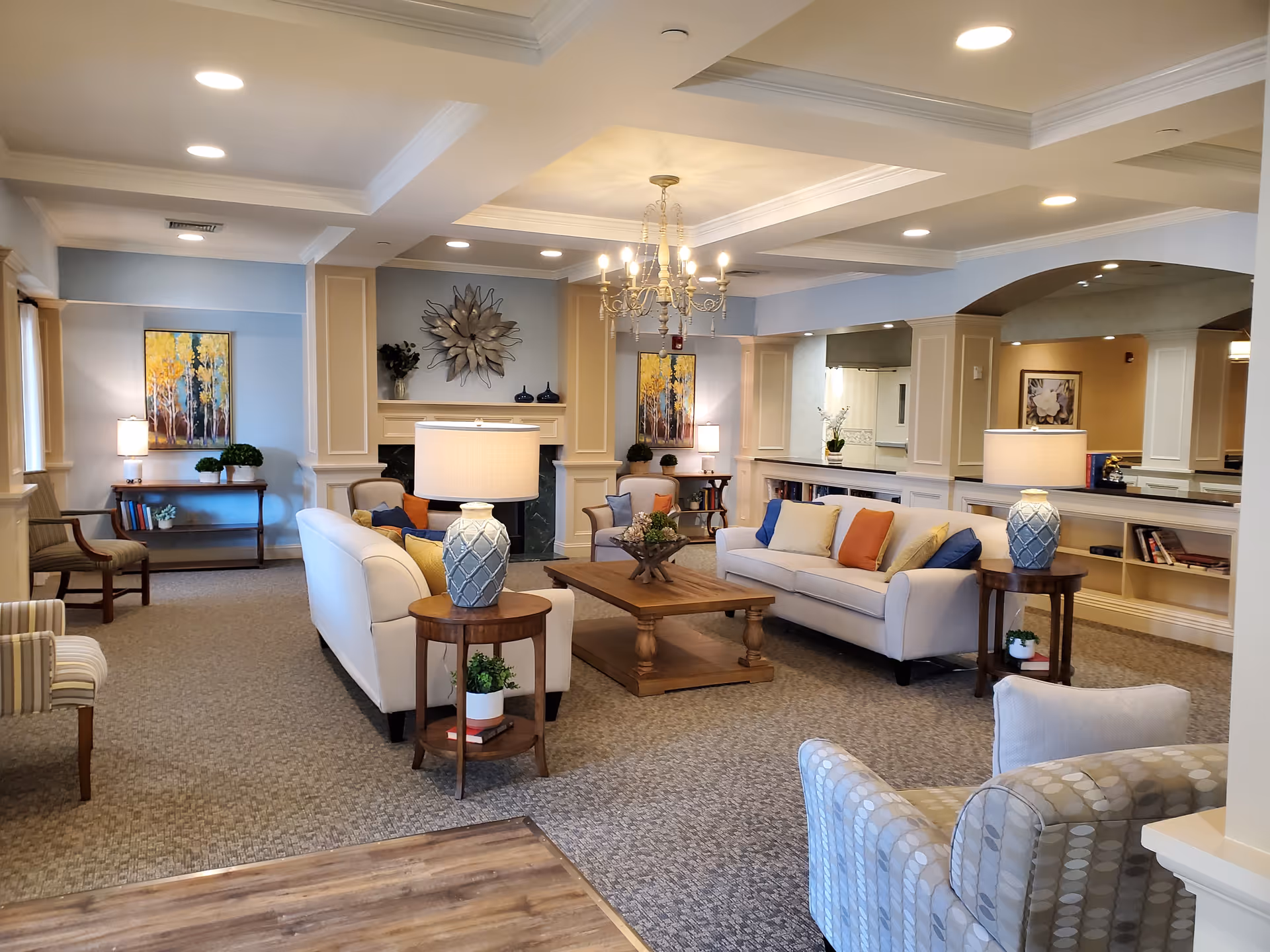A spacious and well-lit living room area with two white sofas adorned with colorful cushions, a wooden coffee table in the center, two side tables with lamps, and additional seating including armchairs. The room features a decorative chandelier, wall art, plants, and built-in shelves with books and decor items. The walls are painted in soft blue and beige tones, and the carpeted floor adds warmth to the space.