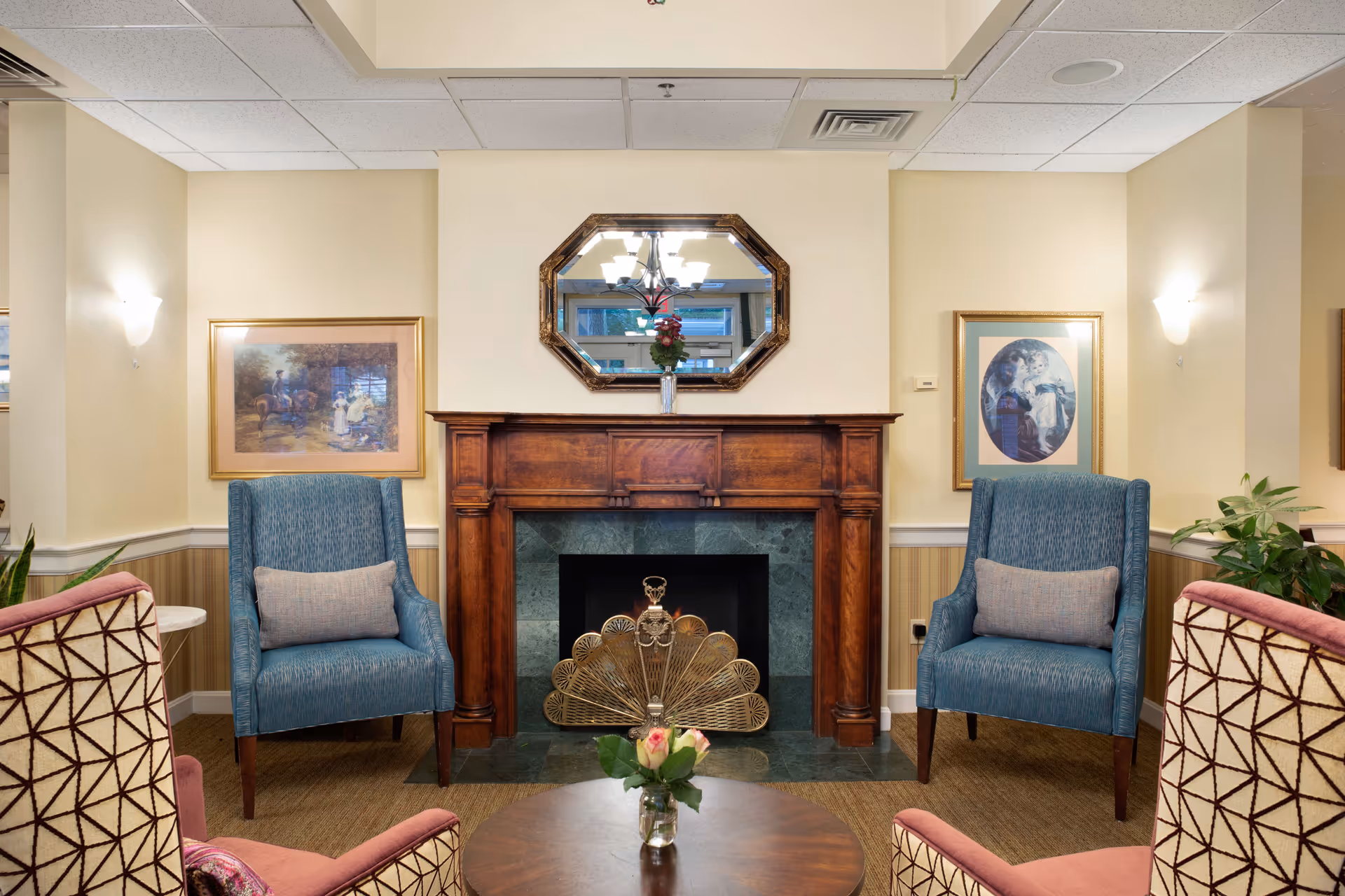 Cozy common-room seating area with a wooden fireplace mantel, an octagonal mirror, two blue armchairs, patterned chairs and a coffee table with flowers.