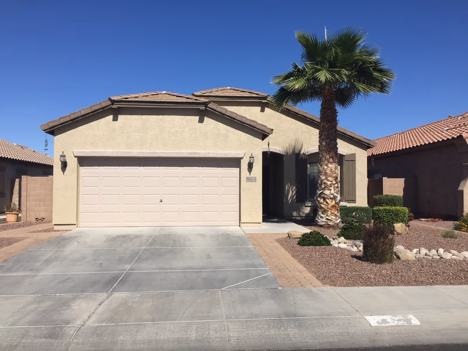 Front exterior view of a single-story beige stucco house with a two-car garage, a palm tree, and desert landscaping including rocks and shrubs under a clear blue sky.