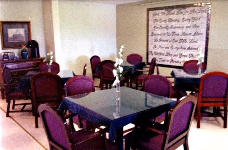 A dining room with multiple square tables covered with dark tablecloths, each table surrounded by purple cushioned chairs. Each table has a small vase with white flowers. On the left side, there is a wooden sideboard with decorative items and a framed picture above it. A large framed piece of calligraphy or text art hangs on the far wall.