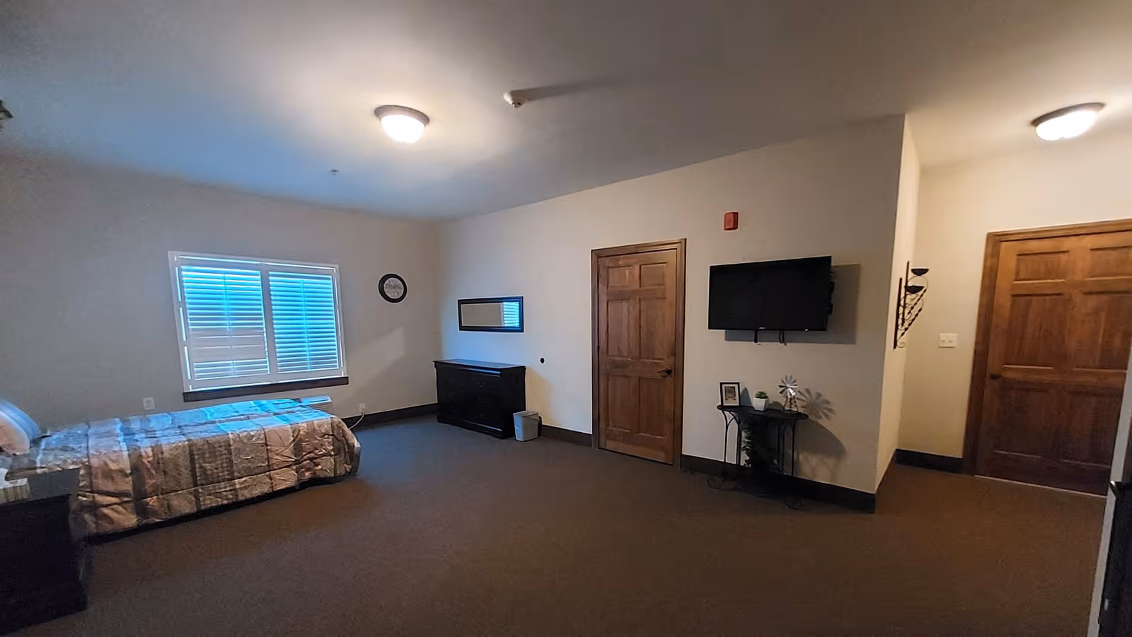 A spacious bedroom in a senior living facility with a bed on the left side, a window with blinds above the bed, a clock on the wall, a dresser with a mirror above it, a small trash bin, a wall-mounted TV, and two wooden doors. The room has carpeted flooring and neutral-colored walls.
