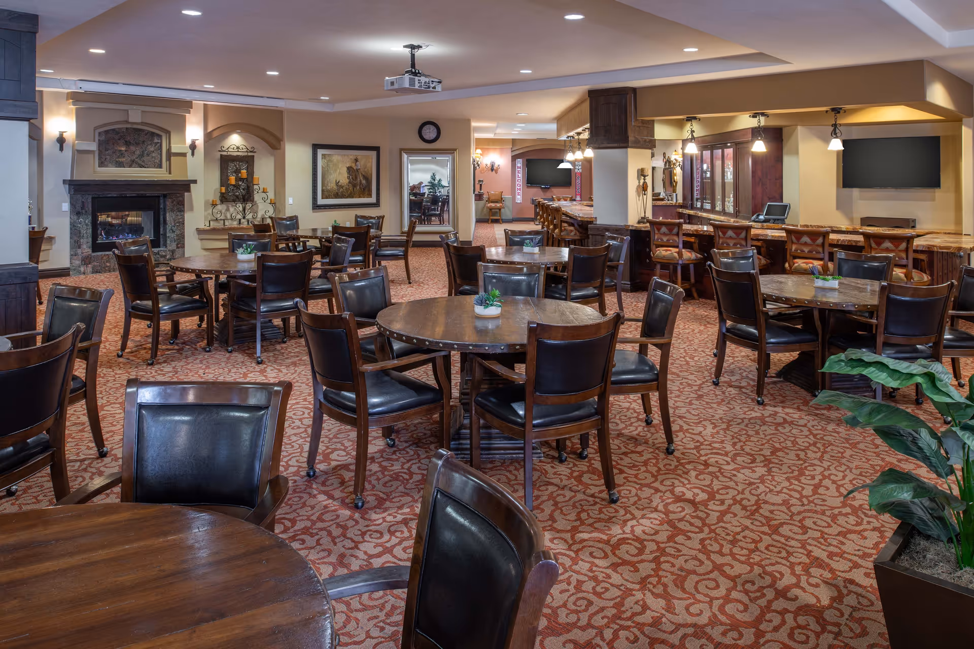 A spacious dining area with multiple round wooden tables and dark leather chairs arranged neatly on a patterned carpet. There is a fireplace on the left wall, framed artwork, and a bar area with high chairs on the right. The room is well-lit with ceiling lights and pendant lamps, and there is a large flat-screen TV mounted on the wall near the bar. A potted plant is visible in the foreground on the right side.
