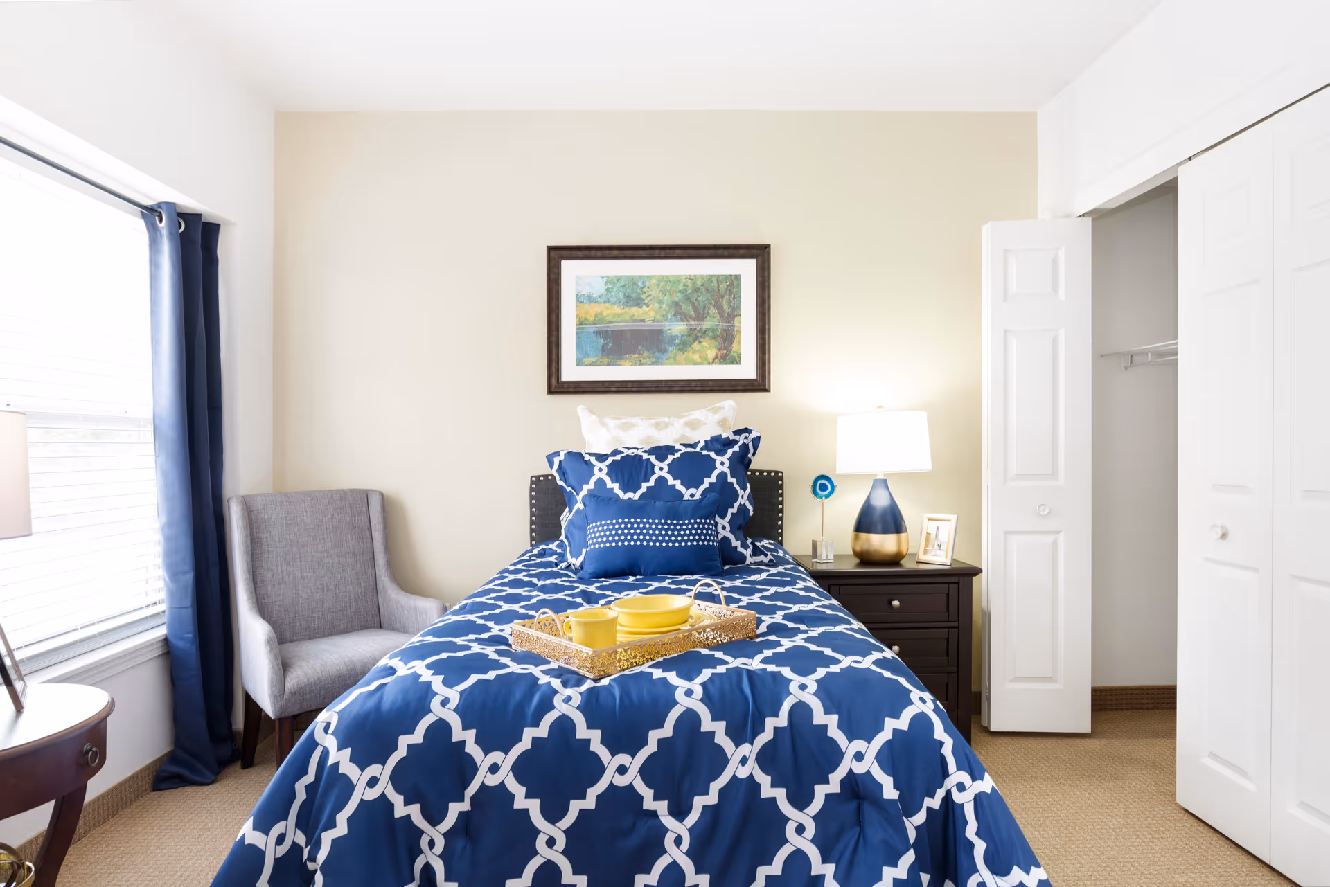 A neatly made single bed with blue and white patterned bedding in a bright bedroom. There is a gray armchair next to a window with blue curtains on the left, a dark wooden nightstand with a lamp, a small framed photo, and a decorative item on the right. Above the bed hangs a framed landscape painting. A closet with white bi-fold doors is partially open on the right side of the room.