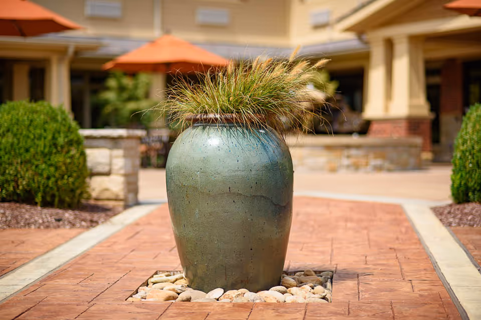 A large green ceramic planter with ornamental grass sits in the center of a paved courtyard area, surrounded by small rocks. In the background, there are trimmed bushes, stone walls, and buildings with orange umbrellas providing shade.