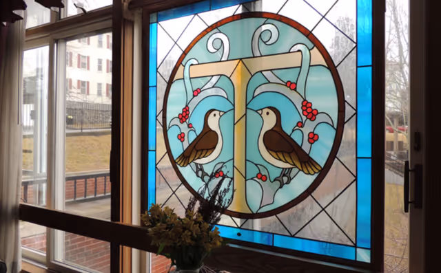 Decorative stained-glass window featuring two birds and floral motifs with a vase of flowers on the windowsill and an outdoor courtyard visible beyond.