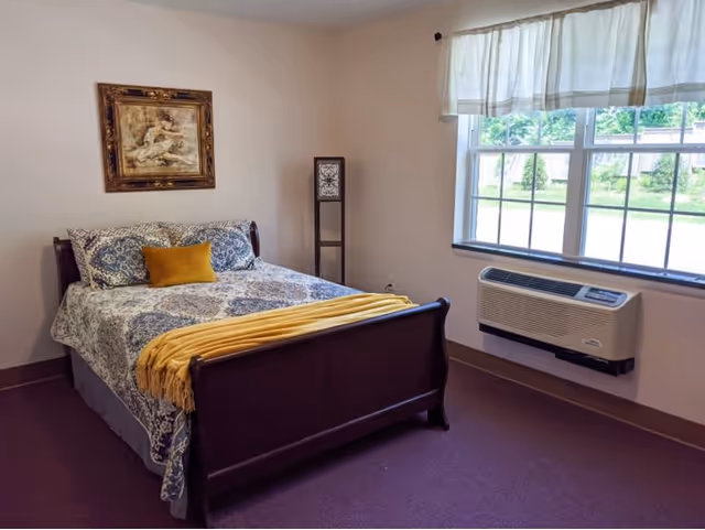 A bedroom with a wooden bed frame, a bed made with patterned blue and white bedding, a mustard yellow throw blanket and pillow, a tall decorative lamp in the corner, a framed artwork on the wall, and a window with white curtains letting in natural light. Below the window is a wall-mounted air conditioning unit.