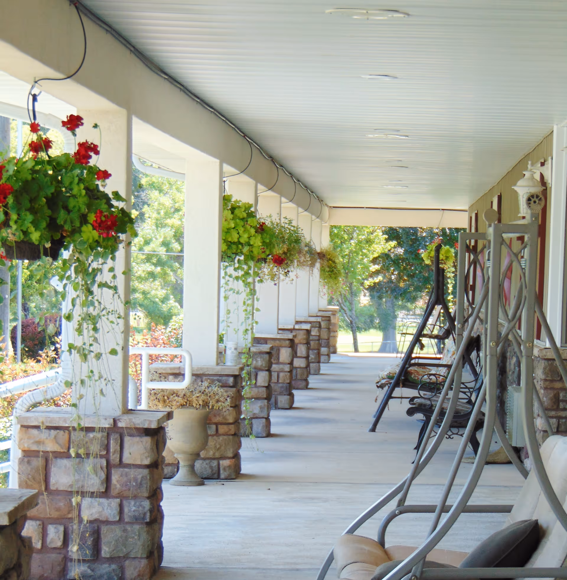 A long covered porch with hanging flower baskets and stone pillars. Several cushioned swing chairs and benches are arranged along the right side, with trees and greenery visible in the background.