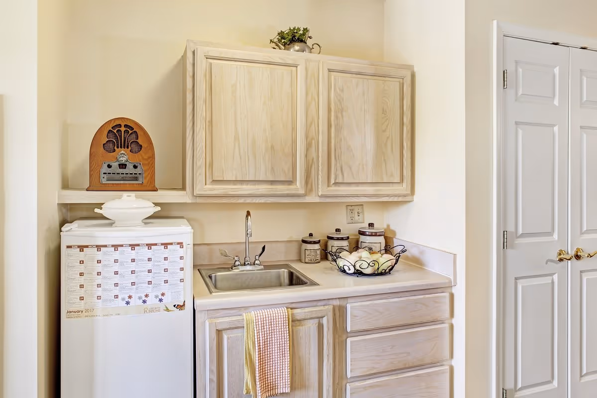 A small kitchen area with light wood cabinets, a stainless steel sink with a faucet, and a countertop holding three ceramic canisters and a wire basket filled with apples. A white mini refrigerator with a calendar on the front is next to the counter, topped with a white ceramic dish and a vintage-style radio. To the right, there is a closed white door with gold handles.