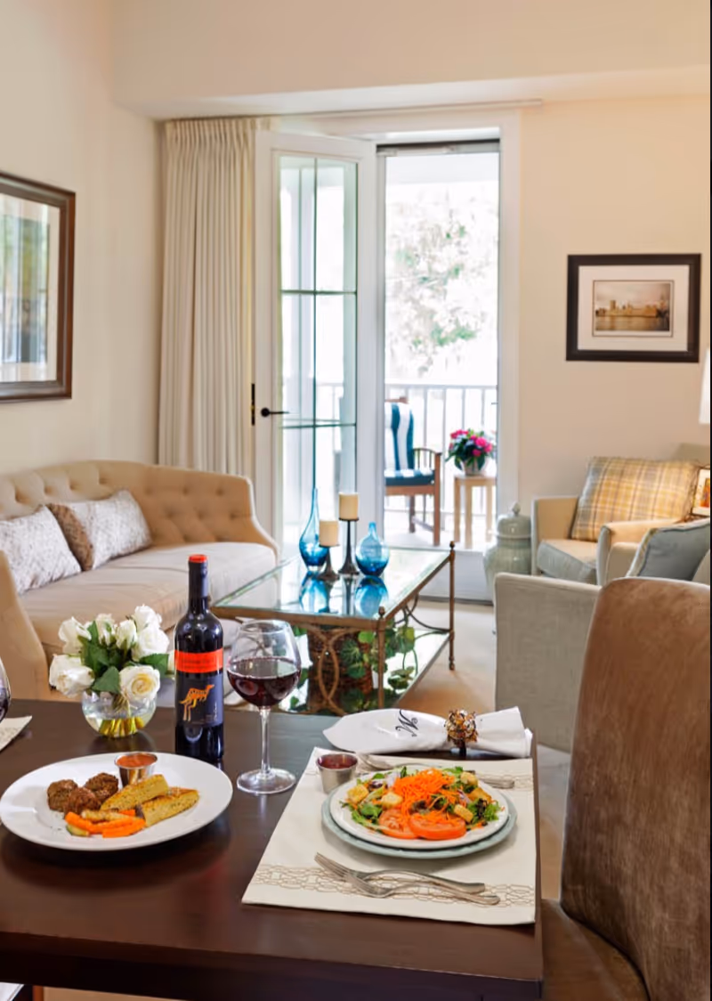 A cozy living room with a beige tufted sofa, armchairs, and a glass coffee table. A dining table in the foreground is set with plates of food, a bottle of red wine, and two glasses of wine. There is a door leading to a balcony with a chair and a small table with flowers outside. The room is decorated with framed pictures and a vase of white roses on the dining table.