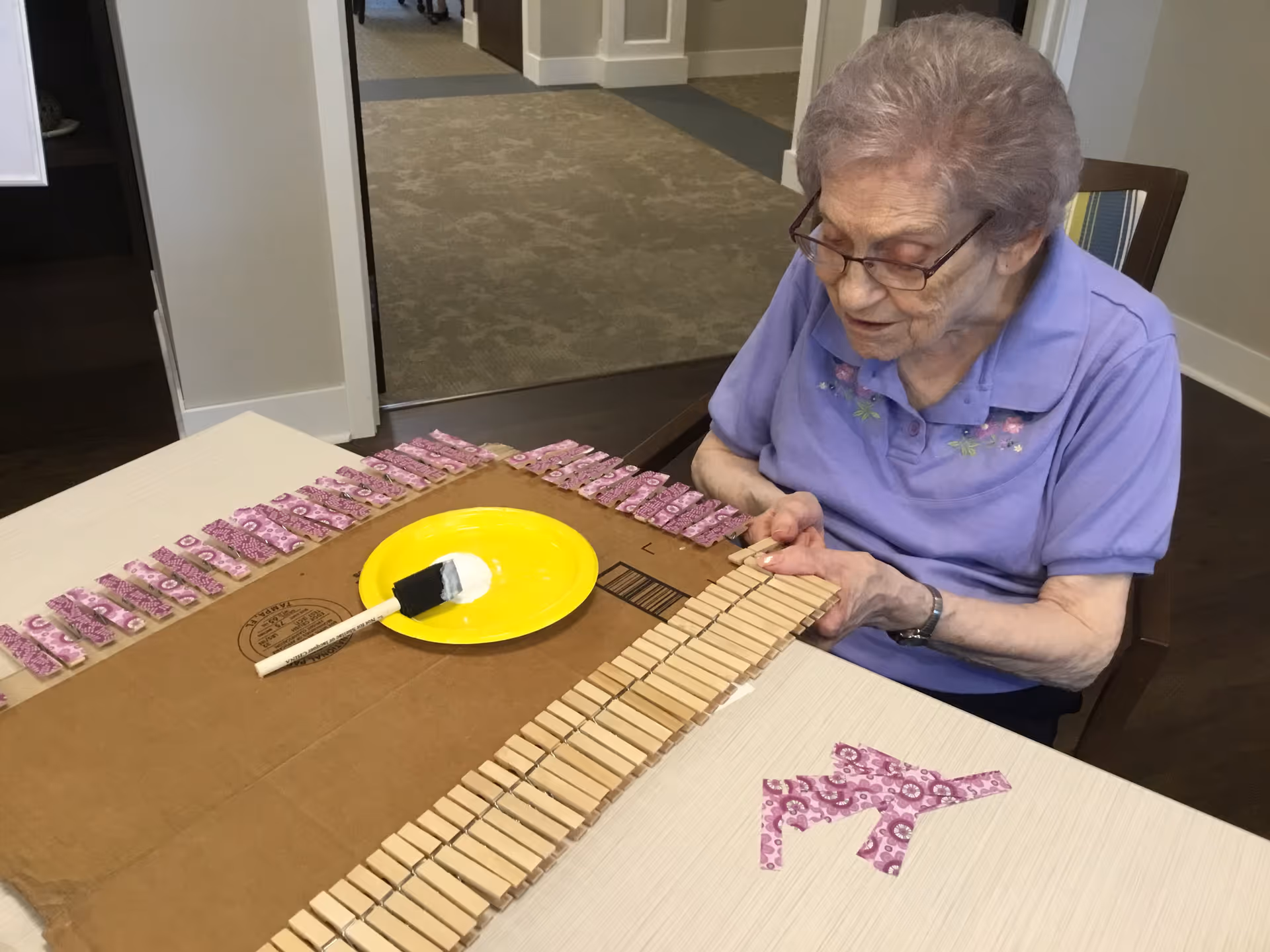 An elderly woman wearing glasses and a purple shirt is sitting at a table, arranging wooden clothespins on a piece of cardboard. There is a yellow plate with a paintbrush on the table, and several clothespins are lined up along the edges of the cardboard.