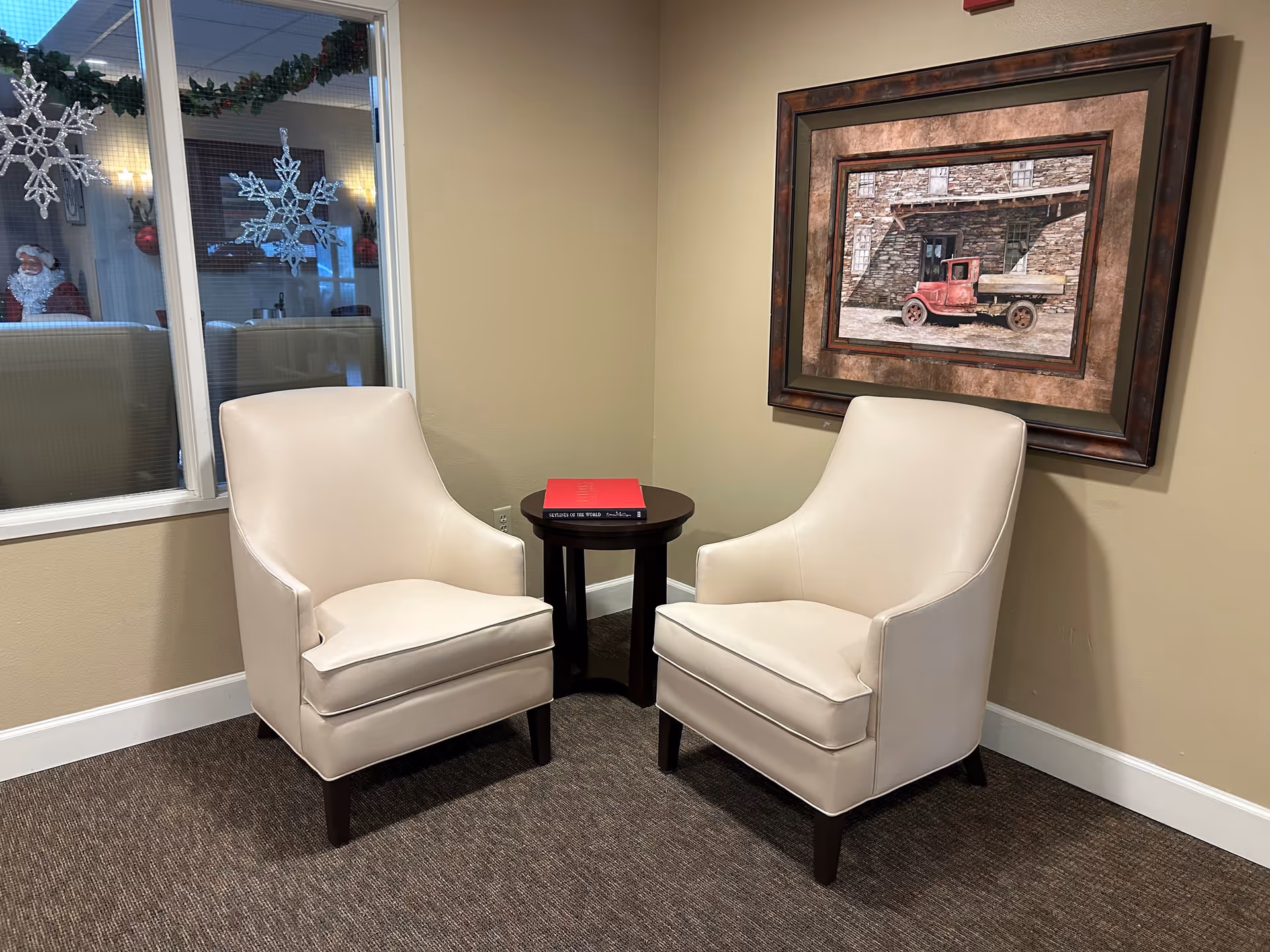 Two cream-colored armchairs positioned around a small round dark wooden table with a red book on it, set against a beige wall with a framed picture of a vintage red truck. A window to the left shows a reflection of holiday decorations including snowflakes and a Santa Claus figure.