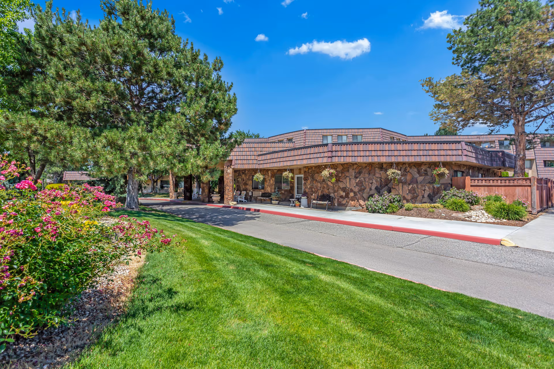 Front entrance of a stone-clad senior living community with a covered driveway, green lawn, trees, and landscaping under a blue sky.