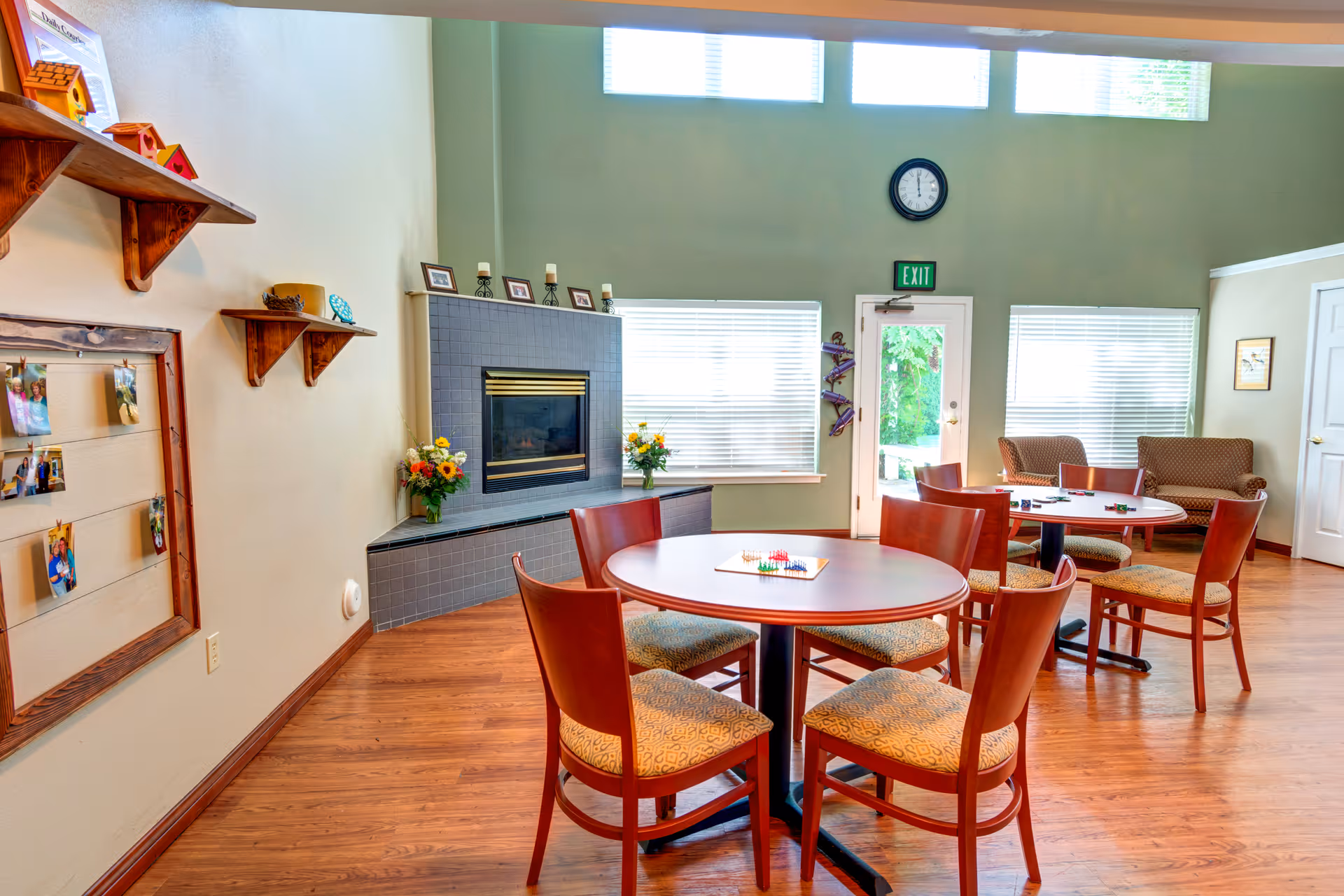 A bright and spacious common area in an assisted living facility featuring round tables with chairs arranged around them. There is a fireplace with a tiled surround, decorated with framed photos and candles. The room has large windows with blinds, a clock above a door marked with an exit sign, and a cozy seating area with two armchairs. The walls are painted in soft green and beige tones, and the floor is wood.