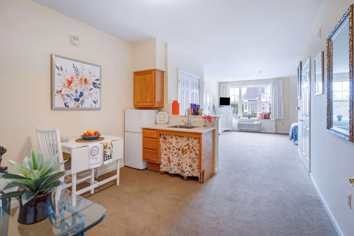 A bright and cozy studio apartment with a small kitchenette featuring wooden cabinets, a white mini fridge, and a sink with a floral curtain underneath. To the left, there is a small white dining table with a chair and a bowl of fruit on top. The room extends into a living and sleeping area with a large window letting in natural light, a white dresser with a TV on top, and a bed partially visible on the right. The walls are light-colored with floral artwork and a large mirror.