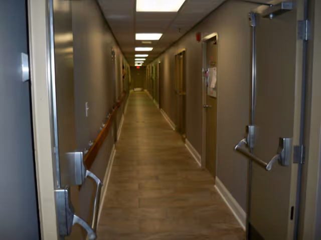 A long, narrow hallway in a facility with beige walls, tiled floor, and multiple closed doors on both sides. The ceiling has recessed lighting panels, and there are handrails along the walls.