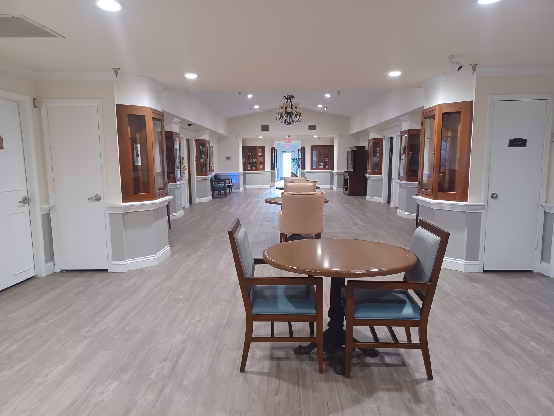 A wide, well-lit communal dining and seating area with round tables and chairs down the center and display cabinets along the sides.