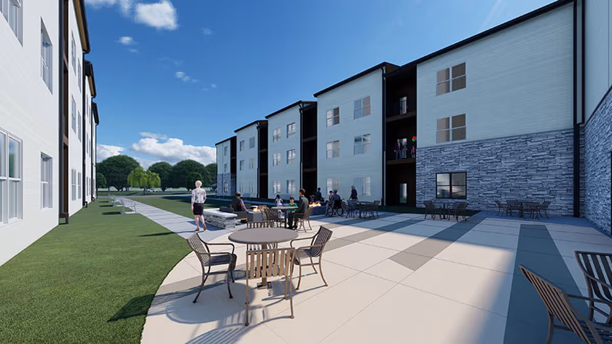 A view of the exterior patio area at Astral at Franklin, featuring outdoor seating, tables, and residents enjoying the space. The building has a modern design with a mix of stone and siding, surrounded by green lawns and trees under a clear blue sky.