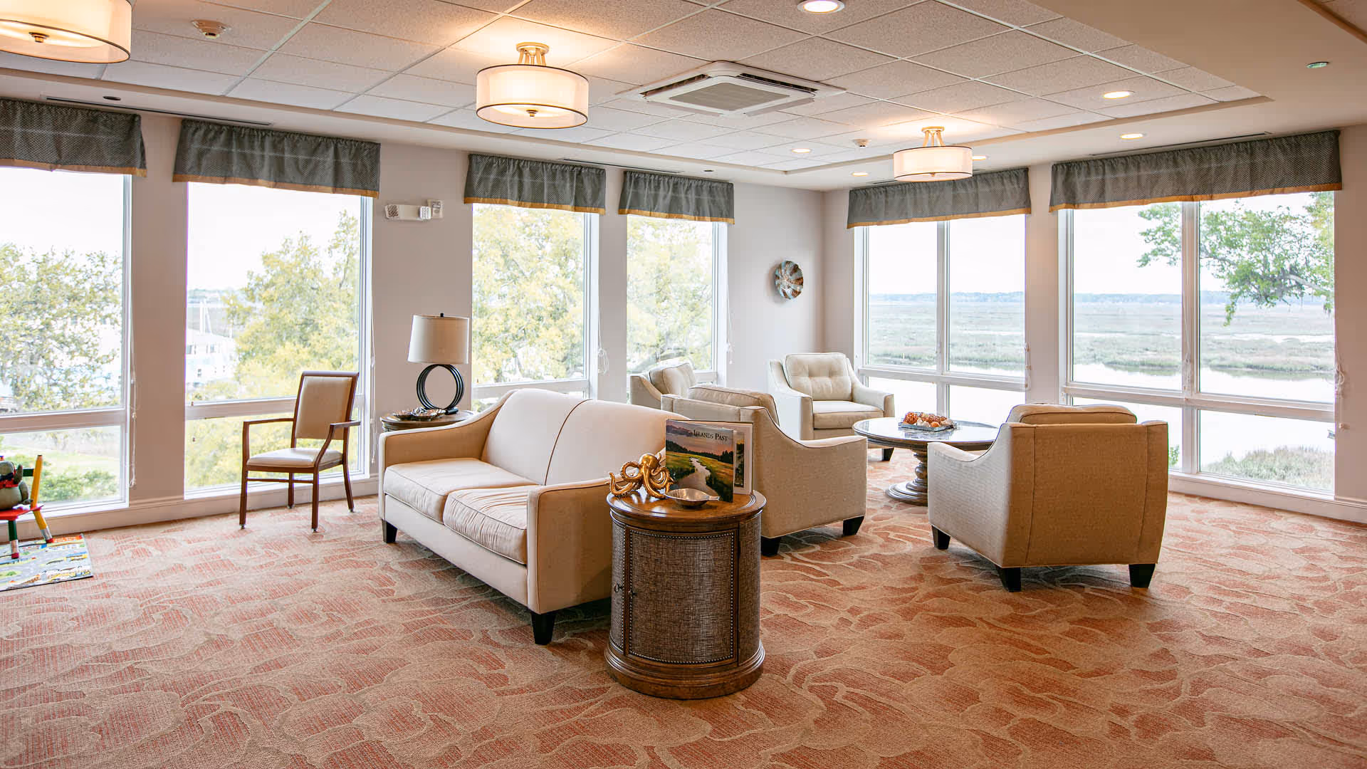 A bright and spacious living room area with large windows offering a scenic view of trees and a water body outside. The room features beige sofas and armchairs arranged around a round coffee table, a side table with a lamp and decorative items, and a patterned carpet. The ceiling has recessed lighting and hanging light fixtures.