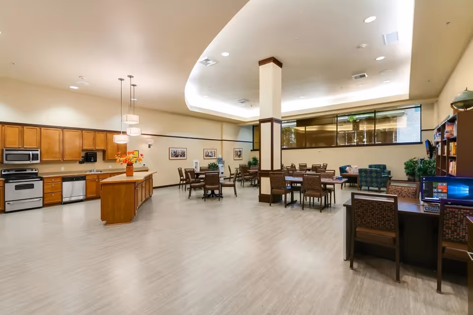 A spacious common area in a senior living facility featuring a kitchen with wooden cabinets, stainless steel appliances, and an island with a vase of flowers. The room includes multiple tables and chairs for dining or activities, a seating area with armchairs, and a desk with a computer. The ceiling has recessed lighting and a large window lets in natural light.