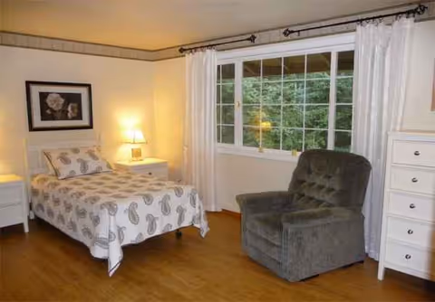 Senior living bedroom featuring a bed, armchair, dresser, and large window.