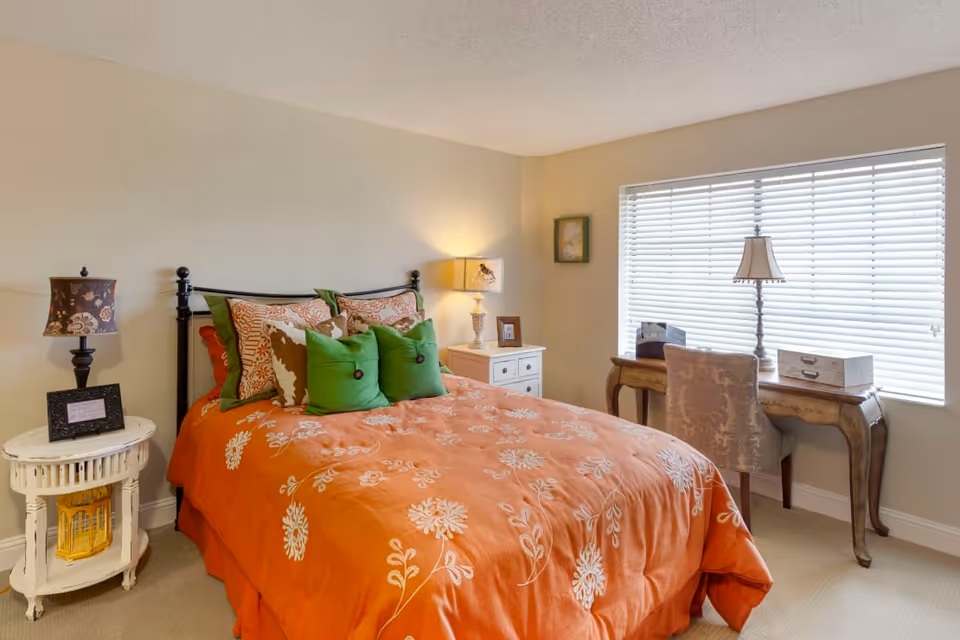A cozy bedroom with a bed covered in an orange floral comforter and multiple pillows in green and brown tones. There are two bedside tables with lamps on each side of the bed. A window with white blinds lets in natural light, and a wooden desk with a chair and a lamp is positioned in front of the window.