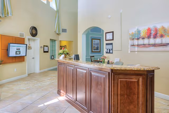 Reception area with a wooden front desk, a granite countertop, and a computer monitor. The room has beige walls, tiled floors, a colorful painting of trees on the wall, and a wall-mounted TV screen displaying information. There are also some chairs and framed pictures in the background.