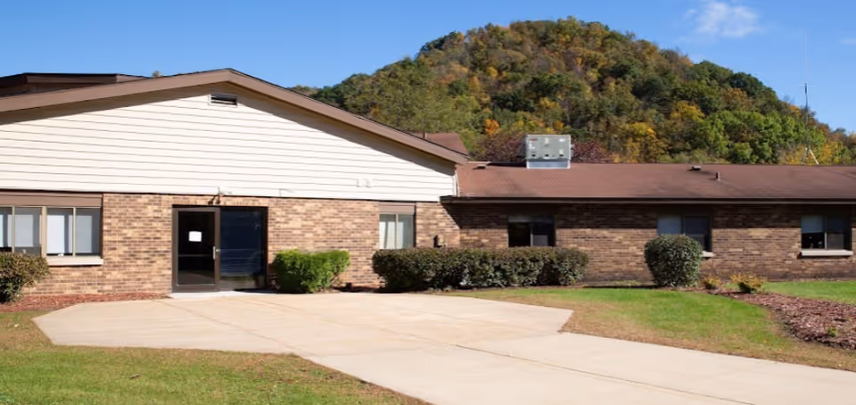 Exterior view of a single-story brick building with a brown roof and a concrete pathway leading to a glass door entrance. The building is surrounded by green grass, bushes, and trees, with a wooded hill in the background under a clear blue sky.