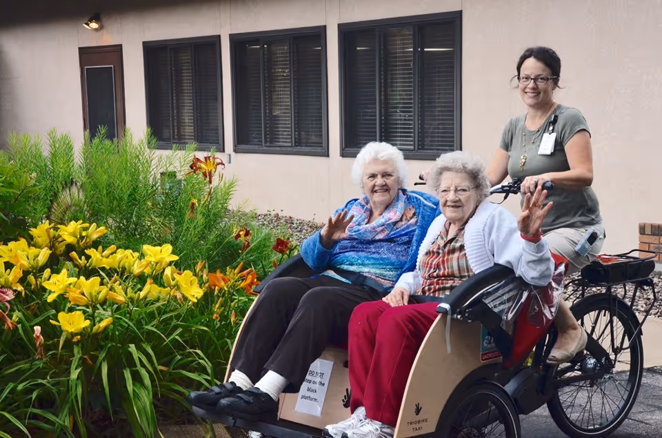 Two elderly women sitting in a tricycle taxi, smiling and waving, with a caregiver riding the bike behind them. They are outside near a building with windows and surrounded by yellow and orange flowers.