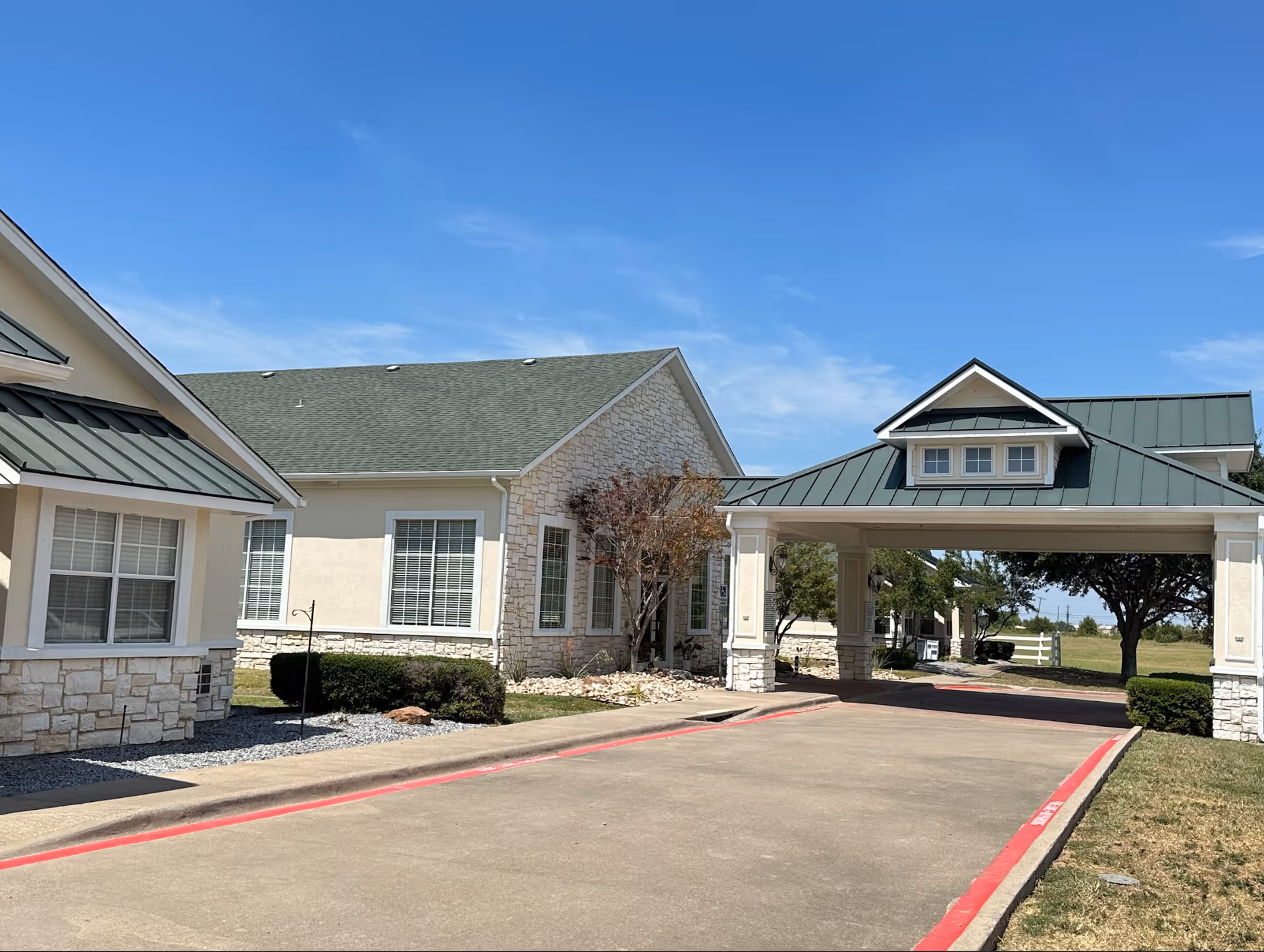Exterior view of Colonial Lodge Assisted Living facility showing a driveway with a covered entrance, beige buildings with stone accents, green roofs, windows with white blinds, and some landscaping including bushes and trees under a clear blue sky.