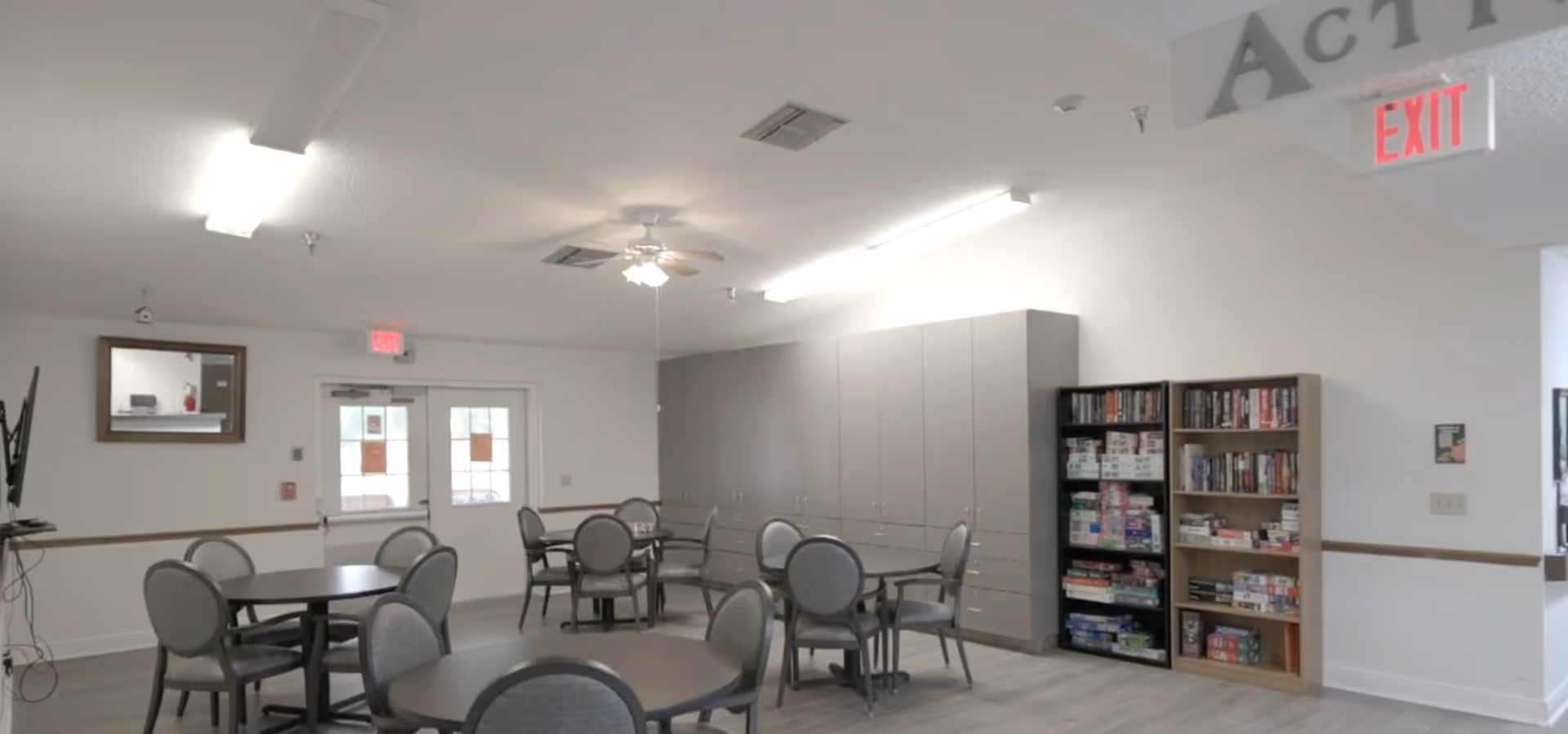 Activity room with round tables and chairs, wall cabinets and shelving filled with board games.