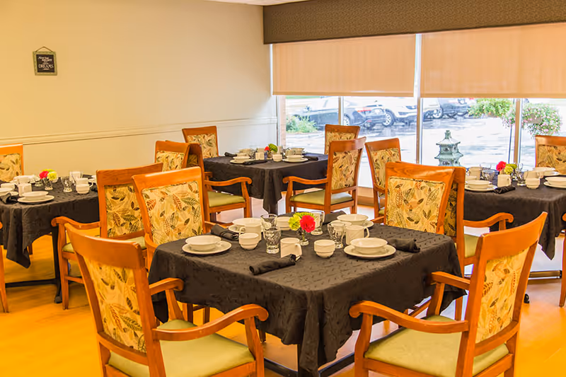 A dining room with several wooden tables covered with black tablecloths, each set with white plates, bowls, cups, and glasses. The chairs have wooden frames with patterned upholstery. Large windows with beige blinds allow natural light to enter, and outside cars and greenery are visible.