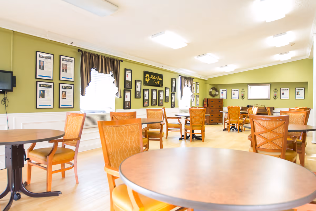 Bright communal dining room with round tables and wooden chairs, green walls decorated with framed photos and a small 'Café' sign.
