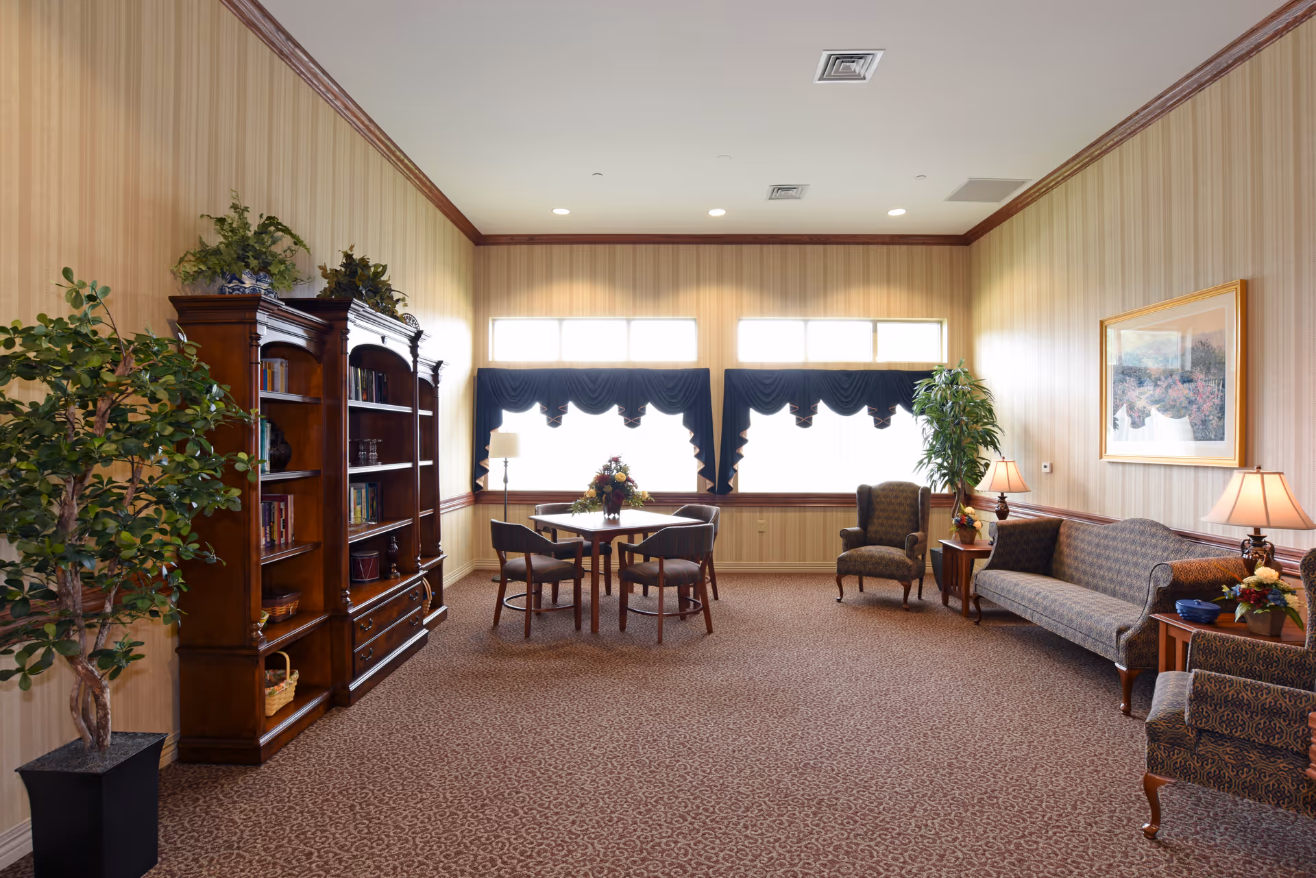 A cozy living room area with patterned carpet and wallpaper, featuring a wooden bookshelf filled with books and decorative items on the left. In the center, there is a small table with four chairs and a floral centerpiece. The room has large windows with dark blue valances letting in natural light. On the right side, there are two upholstered armchairs, a sofa, side tables with lamps, and a framed painting on the wall. Potted plants add greenery to the space.