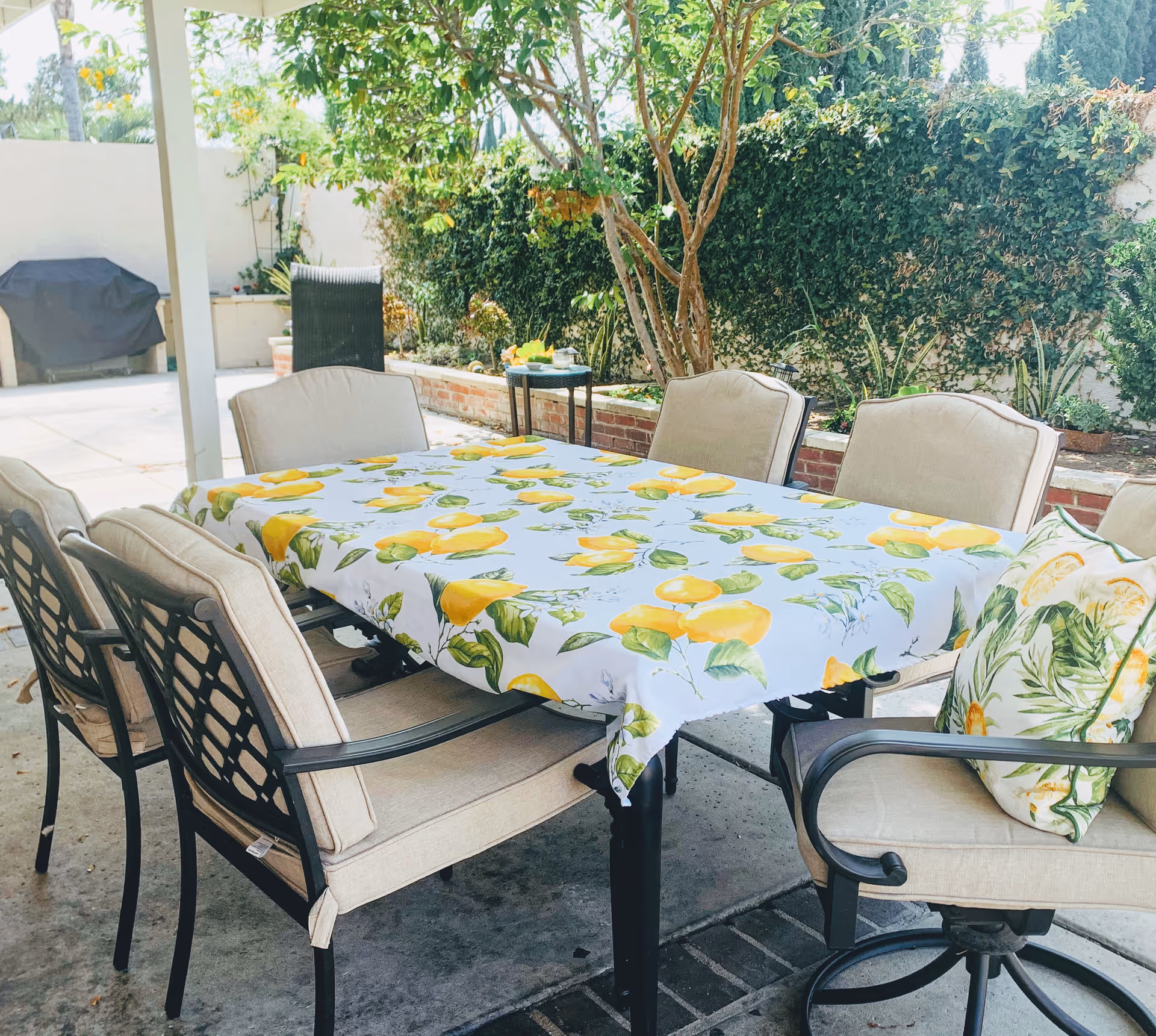 Outdoor patio area with a rectangular table covered with a white tablecloth featuring yellow lemon patterns. The table is surrounded by six cushioned chairs, one with a decorative pillow that has a lemon and leaf design. In the background, there is a barbecue grill covered with a black cover, a small side table, and a garden with green plants and a tree against a white wall.