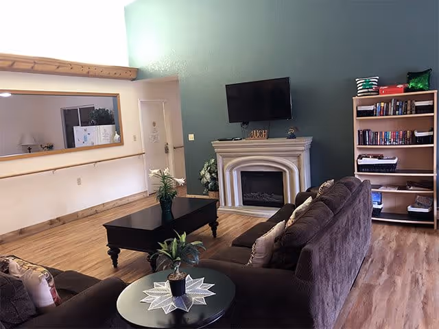 A cozy living room with brown sofas arranged around a black coffee table and a small round side table with a plant. There is a white fireplace with a TV mounted above it on a green accent wall. To the right, there is a wooden bookshelf filled with books and baskets. The room has wooden flooring and a large mirror on the left wall reflecting part of the room.