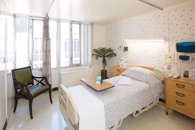 A small, clean hospital-style bedroom with a single bed covered in a light gray patterned blanket. There is a wooden overbed table on the bed, a green cushioned wooden chair near the window, a potted plant in the corner, and two wooden bedside cabinets with medical equipment and personal items. The room has large windows with vertical blinds and patterned wallpaper.