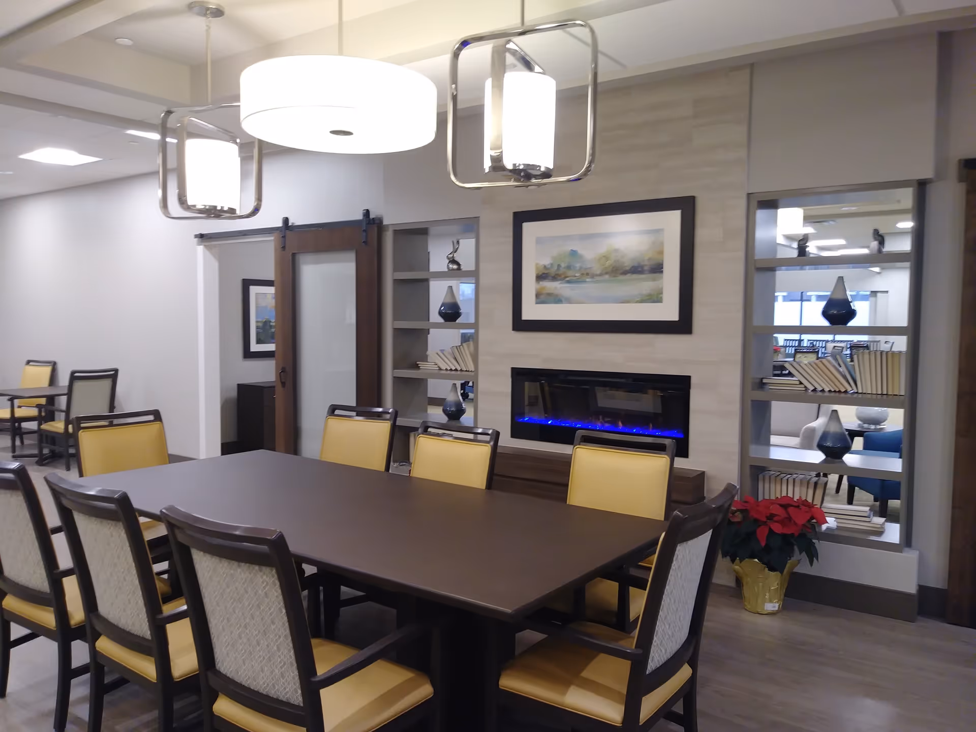 A modern dining room with a large dark wooden table surrounded by eight chairs with yellow cushions. Above the table hang two contemporary light fixtures. On the wall is a built-in electric fireplace with blue flames, above which hangs a framed landscape painting. To the right of the fireplace is a shelving unit with decorative vases and books. A sliding barn door is visible in the background, leading to another room with additional tables and chairs.