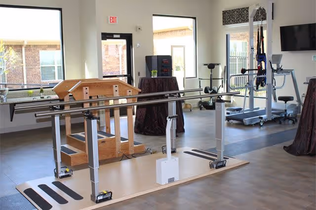 A rehabilitation room with parallel bars for walking exercises, a wooden balance beam structure, exercise equipment including a treadmill, and large windows letting in natural light.