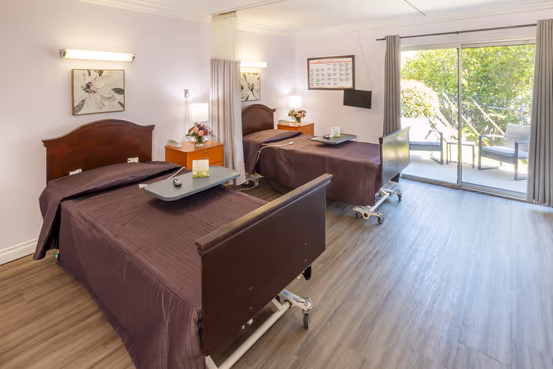 A bright and clean room with two hospital-style beds covered in dark brown bedspreads. Each bed has a wooden headboard and footboard, and a bedside table with a lamp and flower arrangement. There is a wall-mounted TV between the beds and a large sliding glass door leading to an outdoor patio with chairs and greenery visible outside. The room has light-colored walls and wood-look flooring.